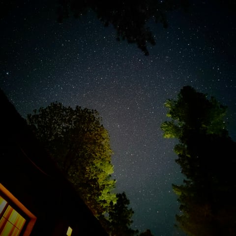 A starry sky with the Milky Way overhead, trees framing the view and a warm cabin window in the corner